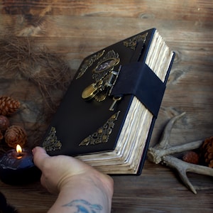 May include: A black leather-bound journal with ornate brass corner accents and a clasp closure. The journal has aged, yellowed pages and a black strap to keep it closed. A key and a lock are attached to the clasp.