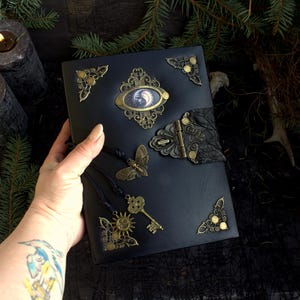 May include: Black leather journal with ornate brass embellishments. Features a central oval design with a moon image, a butterfly, key, and sun charm. Decorative corner pieces and a leaf-shaped clasp add to the vintage aesthetic.