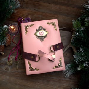 May include: A pink leather journal with a heart-shaped emblem and decorative corner accents. The journal has a dark brown leather strap closure and a key charm. The background includes a lit candle, pine needles, and a wooden surface.