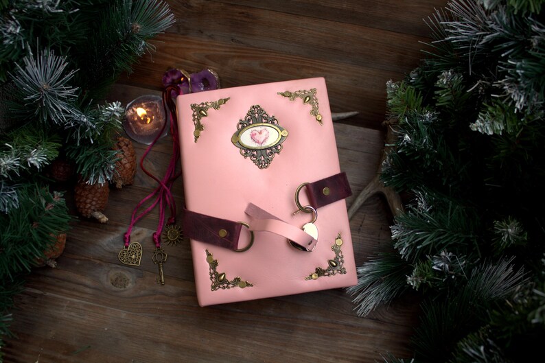 May include: A pink leather journal with a heart-shaped emblem and decorative corner accents. The journal has a dark brown leather strap closure and a key charm. The background includes a lit candle, pine needles, and a wooden surface.