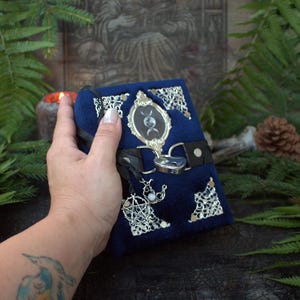 Puede incluir: Un diario pequeño y rectangular cubierto de terciopelo azul oscuro, con esquinas de metal plateado, un marco ovalado central con un símbolo de triple luna y una correa de cuero negro cerrada con un candado plateado en forma de corazón.
