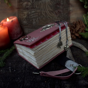 Op de afbeelding: Een oudroze dagboek met een zilveren metalen sluiting en decoratieve zilveren accenten op de kaft. Het boek heeft crèmekleurige pagina's, twee grijze lintbladwijzers met zilveren boombedels en een bijpassende roze leren sluitingsriem.