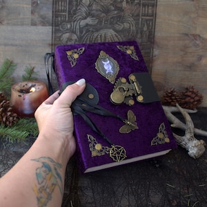 May include: A purple velvet-covered journal featuring ornate brass corner accents, a central oval cabochon with a triple moon symbol, a brass latch closure, and a dangling moth charm along with a pentagram charm attached by black ribbons.