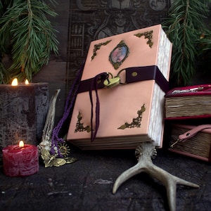 May include: A pink leather-bound book with ornate corner accents and a heart-shaped lock. A dark purple strap secures the book, and a matching necklace with a butterfly pendant lies nearby. The book is surrounded by candles and decorative elements.