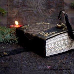 May include: A handcrafted, leather-bound journal with ornate brass corner accents and a decorative clasp. The journal features a textured, dark brown and black cover, aged paper pages, and a black leather strap with a brass ring. The journal is surrounded by pine branches and a lit candle.