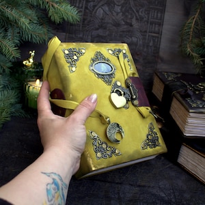 May include: A handcrafted, yellow leather journal with ornate metal corner accents and a heart-shaped lock. The journal features a decorative pendant and a burgundy closure strap. Additional journals are visible in the background.