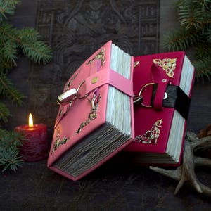 May include: Two leather-bound journals, one pink and one magenta, with ornate gold corner accents. The pink journal has a matching strap and lock closure. The magenta journal has a black strap closure. A lit red candle and evergreen branches are in the background.