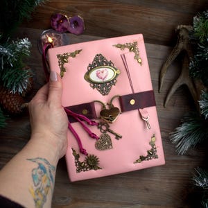 May include: A pink leather journal with ornate brass corner accents and a heart-shaped lock. The journal features a burgundy leather strap, a decorative heart-shaped charm, and a key. The cover has a decorative oval with a heart design.