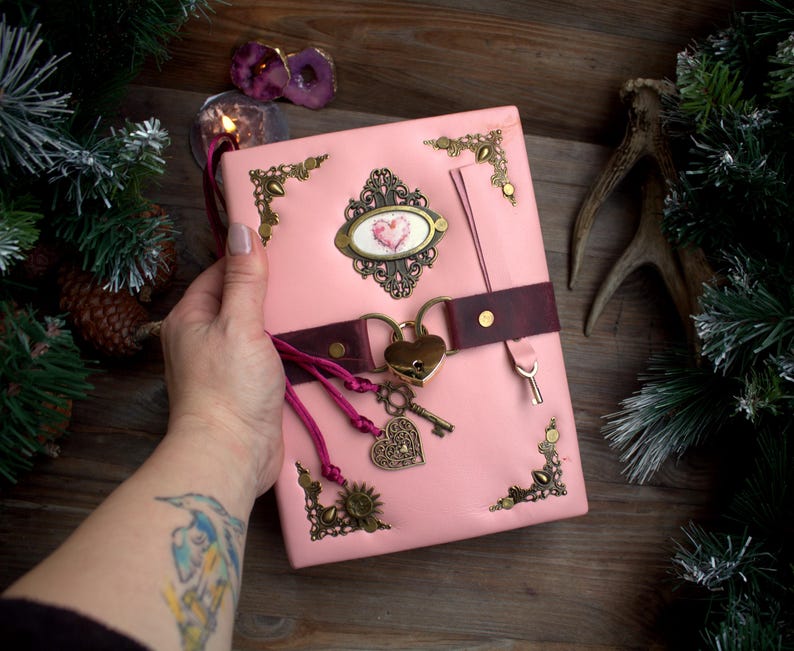 May include: A pink leather journal with ornate brass corner accents and a heart-shaped lock. The journal features a burgundy leather strap, a decorative heart-shaped charm, and a key. The cover has a decorative oval with a heart design.