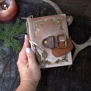 May include: A rectangular journal covered in soft, dusty pink velvet fabric. The cover features ornate brass corner accents, a brown leather strap with a brass buckle closure, and a dangling brass charm shaped like a skeleton key and a feather.