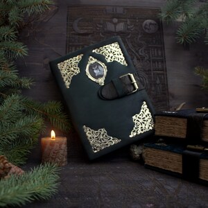 May include: A dark green leather-bound journal with gold filigree corners and a central framed image. The journal features a black leather strap with a buckle closure. A lit candle and evergreen branches are in the foreground.