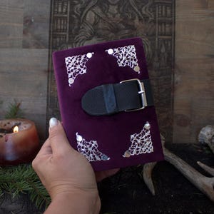 May include: A rectangular journal covered in deep purple velvet fabric. The front cover features four ornate, silver-toned metal corner protectors with intricate filigree patterns. A black leather strap with a silver metal buckle secures the journal.