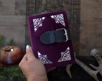 Puede incluir: Un diario rectangular cubierto de tela de terciopelo morado intenso. La portada presenta cuatro protectores de esquina de metal plateado ornamentados con intrincados patrones de filigrana. Una correa de cuero negro con una hebilla de metal plateado asegura el diario.