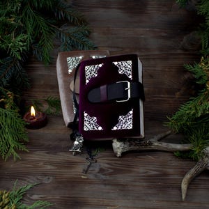 May include: Two closed journals with ornate silver accents and a leather strap. The journal on top is a deep burgundy colour with silver accents. The journal on the bottom is a light brown colour with silver accents. The journals are sitting on a wooden surface with a lit candle and a deer antler.