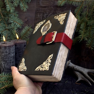 May include: A large, dark green leather-bound book with gold filigree corner accents and a red leather strap with a silver buckle. The book has aged, yellowed pages and a small framed image on the cover.