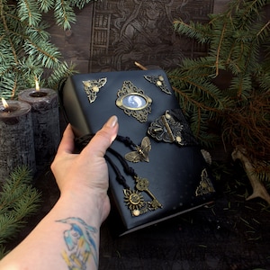 May include: Black leather-bound journal with ornate brass corner pieces, a butterfly clasp, and a decorative pendant with a key and sun charm. The journal features a small oval image and is accented with a black cord.