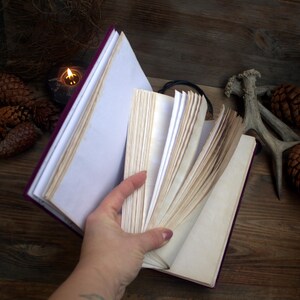 May include: A purple journal with aged, off-white pages is held open, revealing the interior. The cover is a deep purple, and the pages have a vintage, slightly yellowed appearance. The journal is open on a wooden surface.