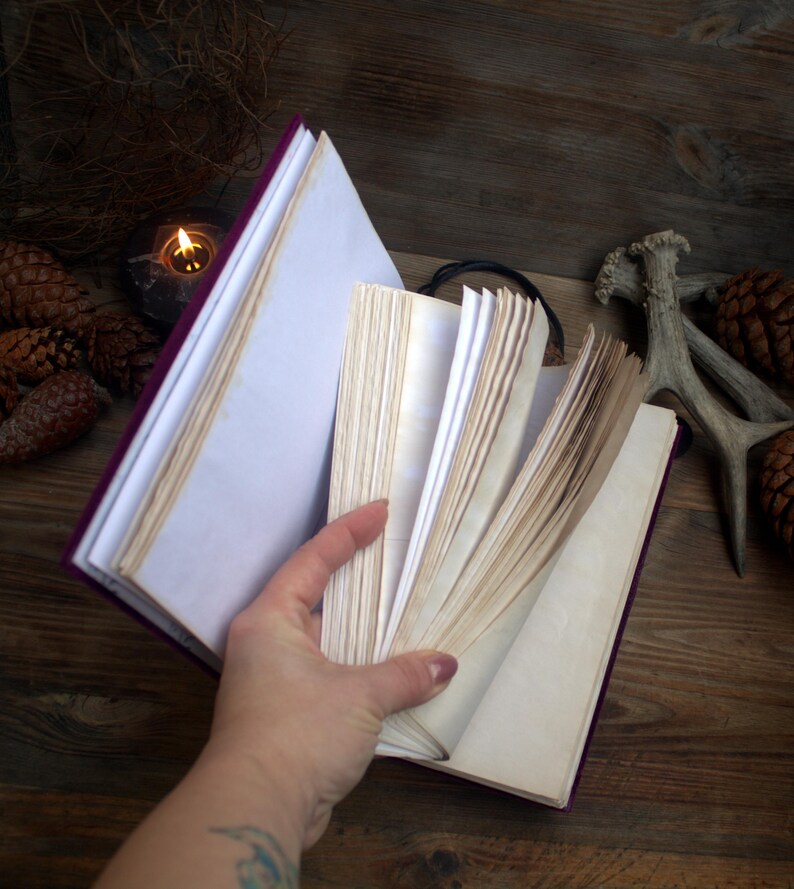 May include: A purple journal with aged, off-white pages is held open, revealing the interior. The cover is a deep purple, and the pages have a vintage, slightly yellowed appearance. The journal is open on a wooden surface.
