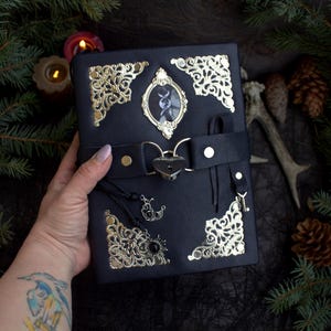 May include: Black leather journal with gold filigree corners and a central oval frame. The journal features a black strap with a heart-shaped lock, and decorative charms. A small image is visible within the oval frame.
