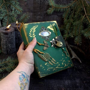 May include: A green journal with gold accents and decorative metal charms, including a butterfly, dreamcatcher, and key. The journal has a leather strap closure and a decorative frame with a small image. The journal is held by a person.