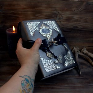 Puede incluir: Un diario de cuero negro con detalles plateados, incluyendo una llave, un dije de mariposa y un cierre de metal decorativo. El diario tiene un símbolo de luna y sol en la parte delantera.