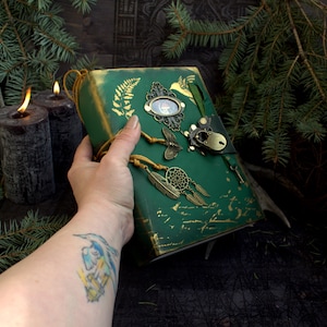 May include: A handcrafted green journal with gold accents, featuring decorative metal charms including a dragonfly, dreamcatcher, and ornate frame. The journal is held in a hand, with a tattoo visible on the wrist. The background includes candles and greenery.