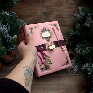 May include: Pink leather journal with a heart-shaped lock and key. The journal features ornate metal corner accents and a decorative oval emblem. A dark brown strap secures the journal, and a pink cord with charms hangs from the side.