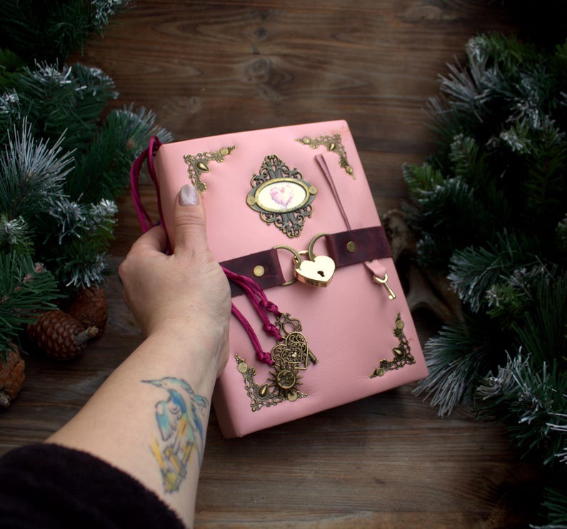 May include: Pink leather journal with a heart-shaped lock and key. The journal features ornate metal corner accents and a decorative oval emblem. A dark brown strap secures the journal, and a pink cord with charms hangs from the side.