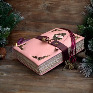 May include: A pink leather-bound journal with ornate corner accents and a burgundy strap closure. The journal features a decorative clasp and a charm with keys, a sun, and leaves. The journal is on a wooden surface.