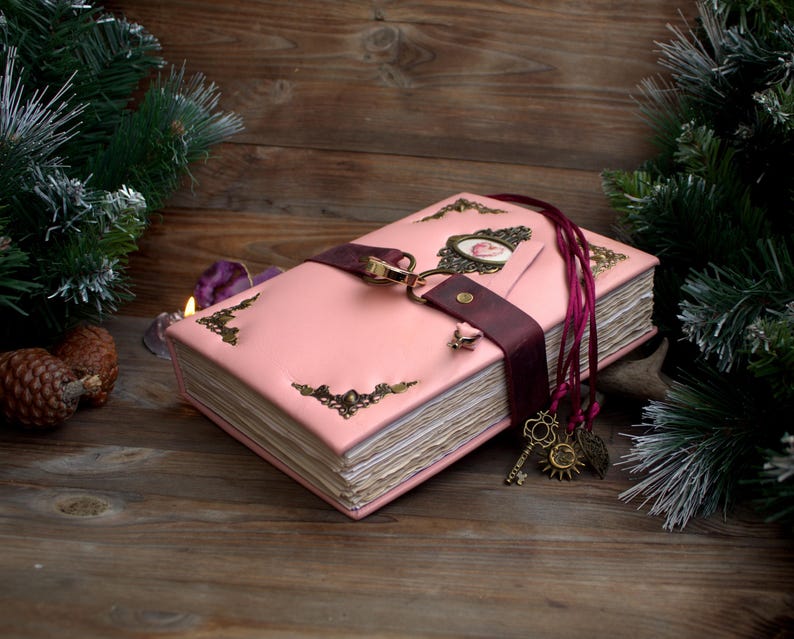 May include: A pink leather-bound journal with ornate corner accents and a burgundy strap closure. The journal features a decorative clasp and a charm with keys, a sun, and leaves. The journal is on a wooden surface.