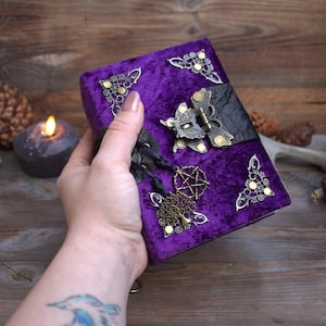May include: A purple velvet journal featuring ornate brass corner protectors, a central brass butterfly-shaped latch, and two black cord charms with a metal pentagram and a tree of life pendant attached to the front cover of the book.