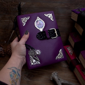 May include: A deep purple journal with silver corner accents and a decorative buckle closure. The journal features a small framed image and a black strap with star-shaped cutouts. A dreamcatcher charm hangs from the journal's spine.
