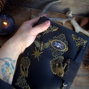 May include: Black leather journal with ornate brass accents, including a moth, star, and dreamcatcher charm. The journal features a decorative lock and a celestial-themed oval emblem. A black ribbon bookmark and key are also visible.