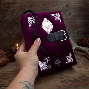 May include: A deep purple velvet journal with silver filigree corners and a black leather strap closure. The journal features a small framed image and a black cord with a key and tree charm.