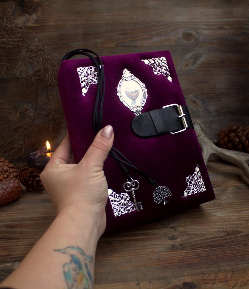 May include: A deep purple velvet journal with silver filigree corners and a black leather strap closure. The journal features a small framed image and a black cord with a key and tree charm.