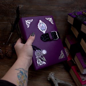 Pode incluir: Um diário de couro roxo com cantos prateados e um fecho de fivela decorativo. O diário apresenta uma pequena imagem emoldurada e um pingente com um apanhador de sonhos, um sol e penas. Diários adicionais estão empilhados ao fundo.