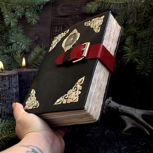May include: A large, black leather-bound book with gold filigree corner accents and a red leather strap closure. The book has a small framed image on the cover and aged, off-white pages. The book is held in a hand with a tattoo.