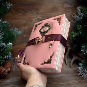 May include: A pink leather journal with ornate corner and center accents. A burgundy strap with a gold clasp secures the journal. A small key charm hangs from the clasp. The journal is held in a hand.