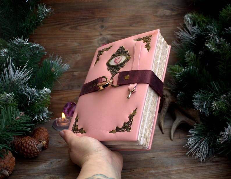 May include: A pink leather journal with ornate corner and center accents. A burgundy strap with a gold clasp secures the journal. A small key charm hangs from the clasp. The journal is held in a hand.