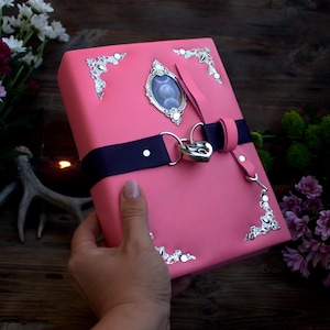 May include: Pink journal with silver corner accents and a decorative oval centerpiece. A navy blue strap secures the journal with a silver heart-shaped lock. A pink leather strap is attached to the lock. The journal is held in a hand.