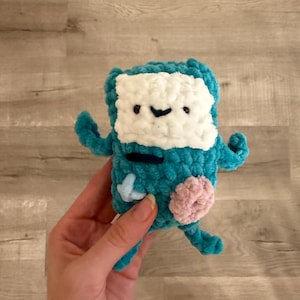 May include: A handmade teal crochet plush toy with a white face, black stitched details, and a pink patch. The toy has small arms and legs, and is held in a hand. The plushie is a character from the animated series Adventure Time.