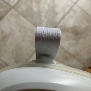 May include: Close-up of a white and gray object with a curved, ribbed handle. The handle is attached to a white, rounded base. The object appears to be a part of a larger item, possibly a container or appliance.