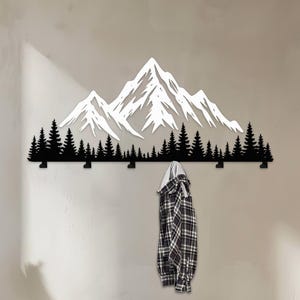 May include: A black metal wall-mounted coat rack featuring a mountain and forest design. The mountain peaks are white, and the trees are black. A plaid shirt hangs from one of the hooks. The rack is mounted on a light-colored wall.