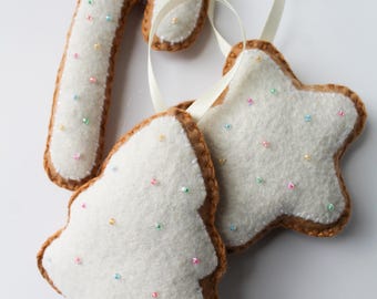 May include: Three felt Christmas ornaments shaped like a candy cane, a star, and a Christmas tree. Each ornament is white with brown edges and decorated with colorful sprinkles.