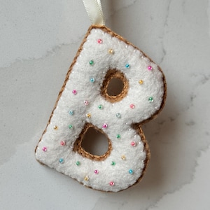 Peut inclure: Ornement en forme de lettre B en feutre blanc avec un contour en feutre marron et des paillettes colorées.