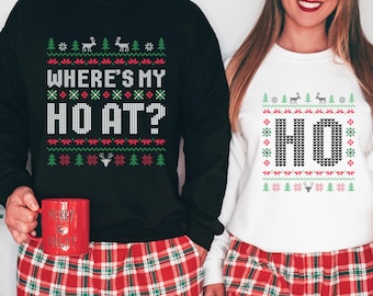 May include: Matching Christmas sweaters, one black and one white, with festive holiday designs. The black sweater says "WHERE'S MY HO AT?" and the white sweater says "HO". Both have reindeer and tree patterns. The models are wearing red plaid pajama pants.