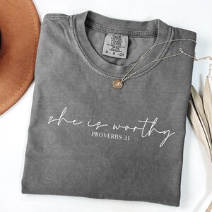 May include: A gray Comfort Colors t-shirt with the words "she is worthy" and "Proverbs 31" printed in white script. A silver necklace with a circular pendant rests on the shirt. A brown hat and dried plant are in the background.