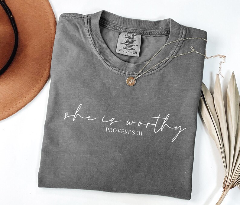 May include: A gray Comfort Colors t-shirt with the words "she is worthy" and "Proverbs 31" printed in white script. A silver necklace with a circular pendant rests on the shirt. A brown hat and dried plant are in the background.