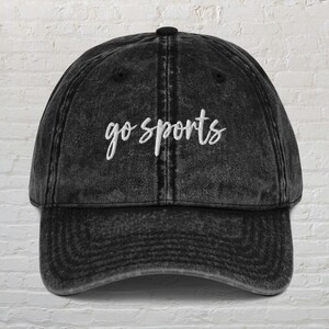 Könnte beinhalten: Eine schwarze, ausgewaschene Baseballkappe mit dem weißen, kursiven Text "go sports" auf der Vorderseite gestickt. Die Kappe hat einen gebogenen Schirm und einen Knopf oben. Der Hut ist aus weichem, abgenutztem Material.