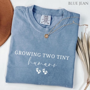 May include: Blue jean colored t-shirt with the text "GROWING TWO TINY humans" in white script. The shirt has a crew neck and short sleeves. A gold necklace with a pendant rests on the shirt.